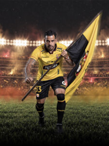 Jugador de Barcelona Sporting Club avanza sosteniendo una bandera amarilla y negra, con el Estadio Banco Pichincha de fondo, previo al partido ante Universidad Católica.